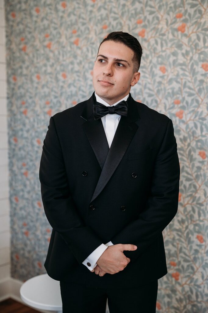 A groom in a black tuxedo poses for a black and white wedding photo.