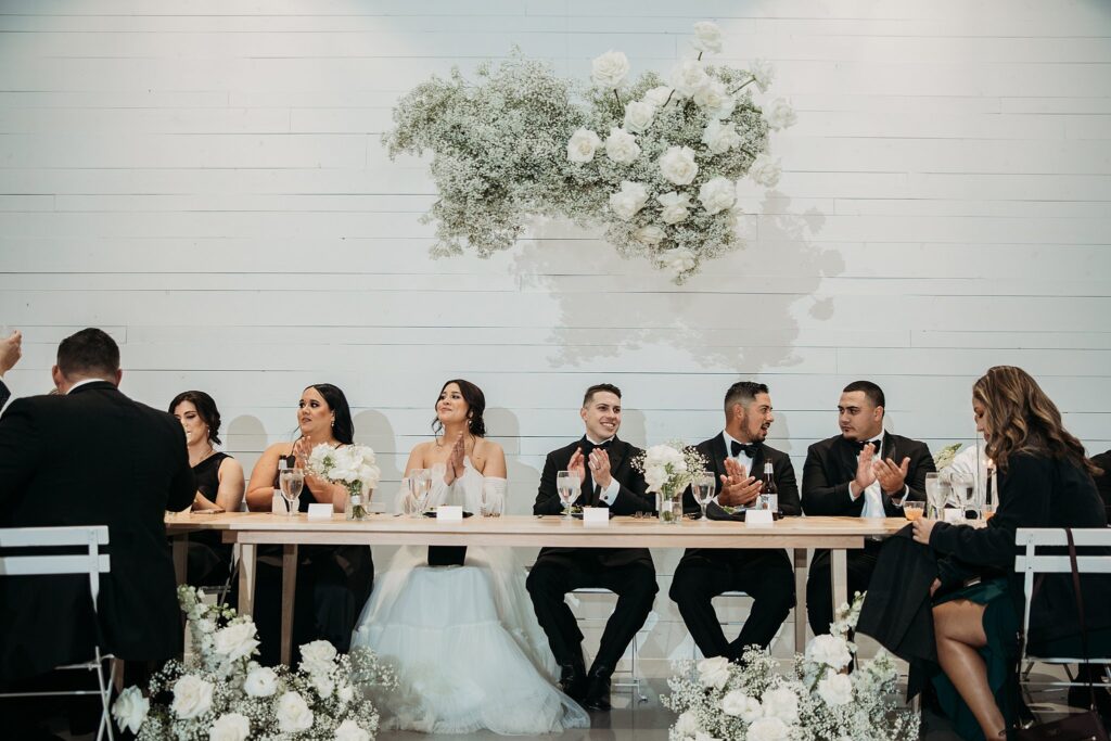 A black and white wedding reception with a bride and groom clapping at their table.