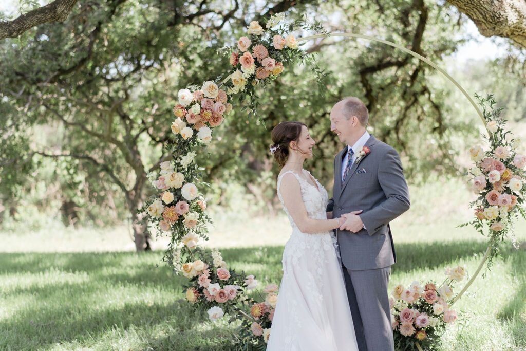A bride and groom standing in front of a circular wedding arch at Pecan Springs Ranch.