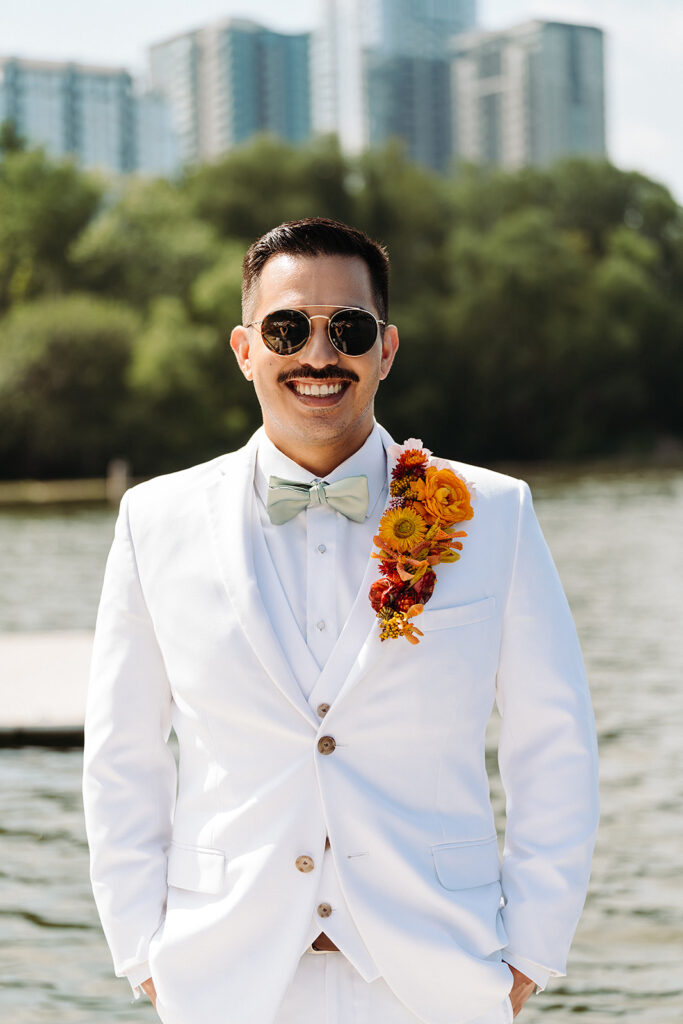 A man in a white suit and sunglasses standing by a body of water.