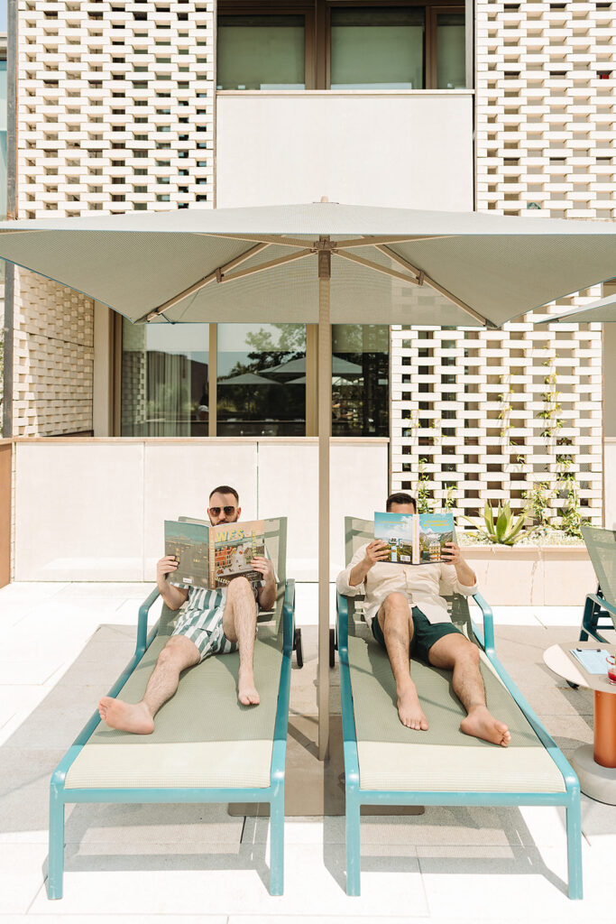 Two men laying on lounge chairs under an umbrella at the South Congress Hotel pool.