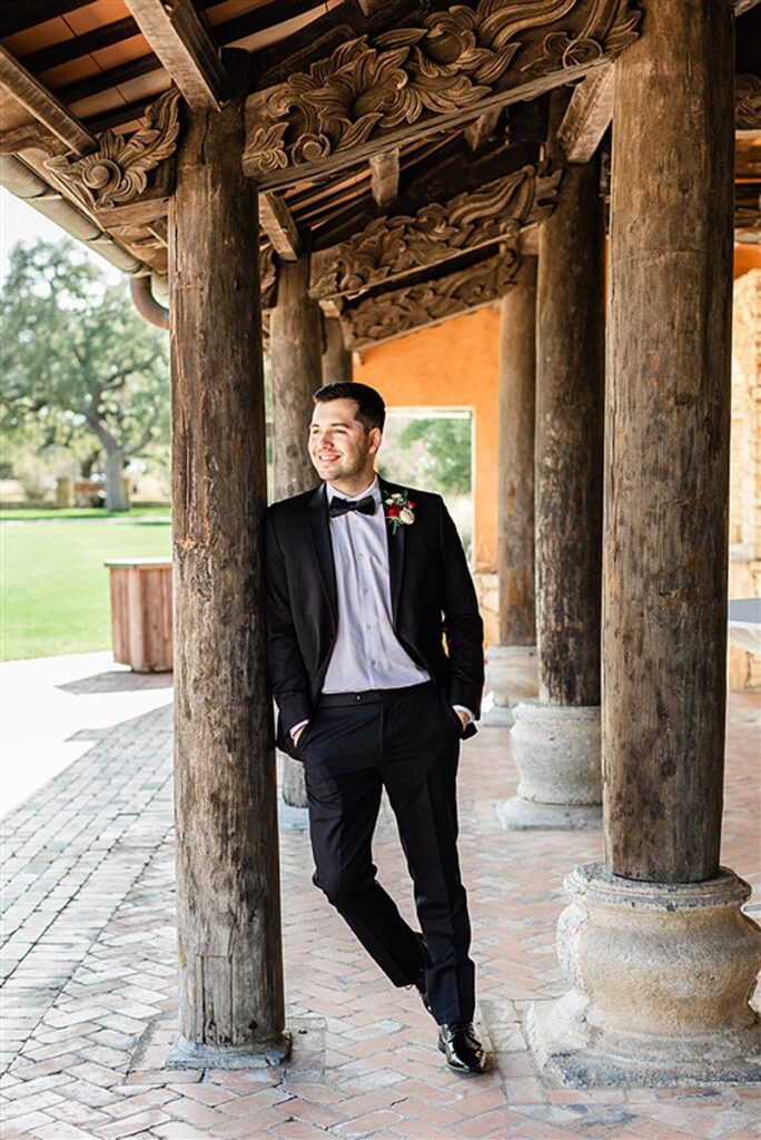 A man in a tuxedo is leaning against a wooden Camp Lucy wedding structure.