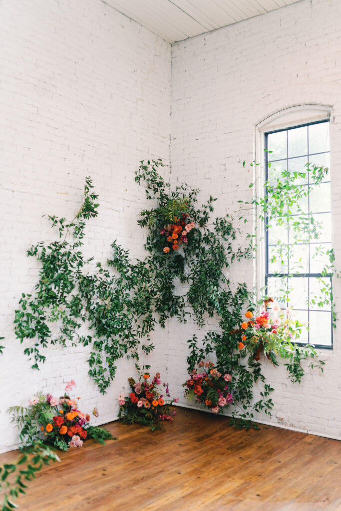 Green leafy vines and colorful flower arrangements decorate a white brick corner at One Eleven East, with a tall window, wood floor, and sunlight streaming in.