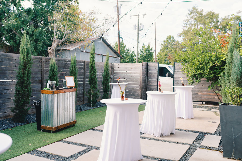 Outdoor event space at One Eleven East with cocktail tables covered in white cloth, a rustic bar setup on the left, string lights overhead, and a wooden fence with trees in the background.