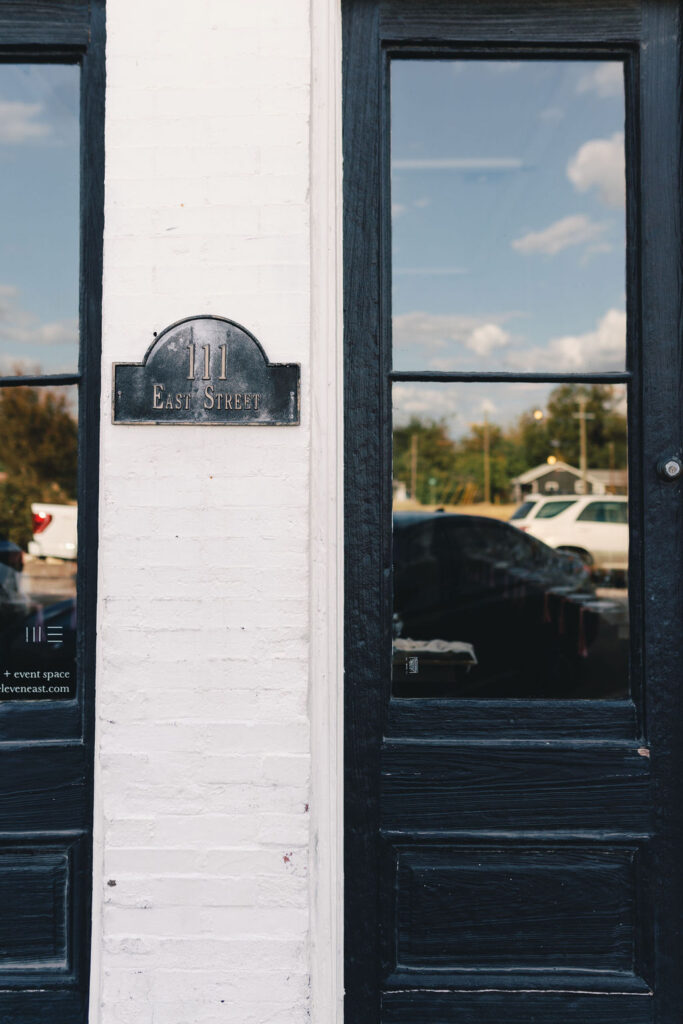 A black metal sign reading “111 East Street” is mounted on a white brick wall between two black-framed glass doors reflecting cars and trees, marking the entrance to One Eleven East.