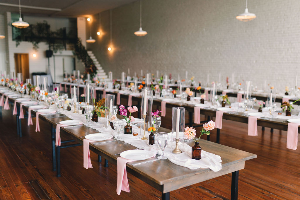 Long wooden tables set for an event at One Eleven East feature white plates, glassware, pink napkins, and small vases of flowers in a modern, airy room with wooden floors and hanging lights.