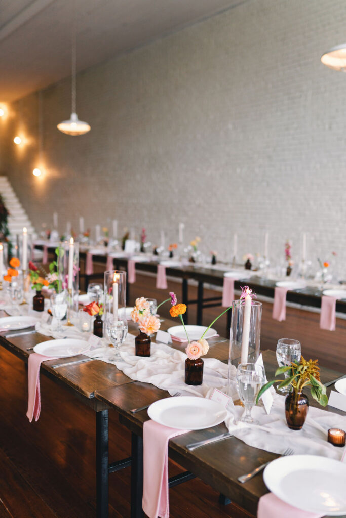 Long wooden tables set for an event with white plates, glassware, pink napkins, candles, and small floral arrangements in brown vases line the airy space at One Eleven East, all against a striking white brick wall.