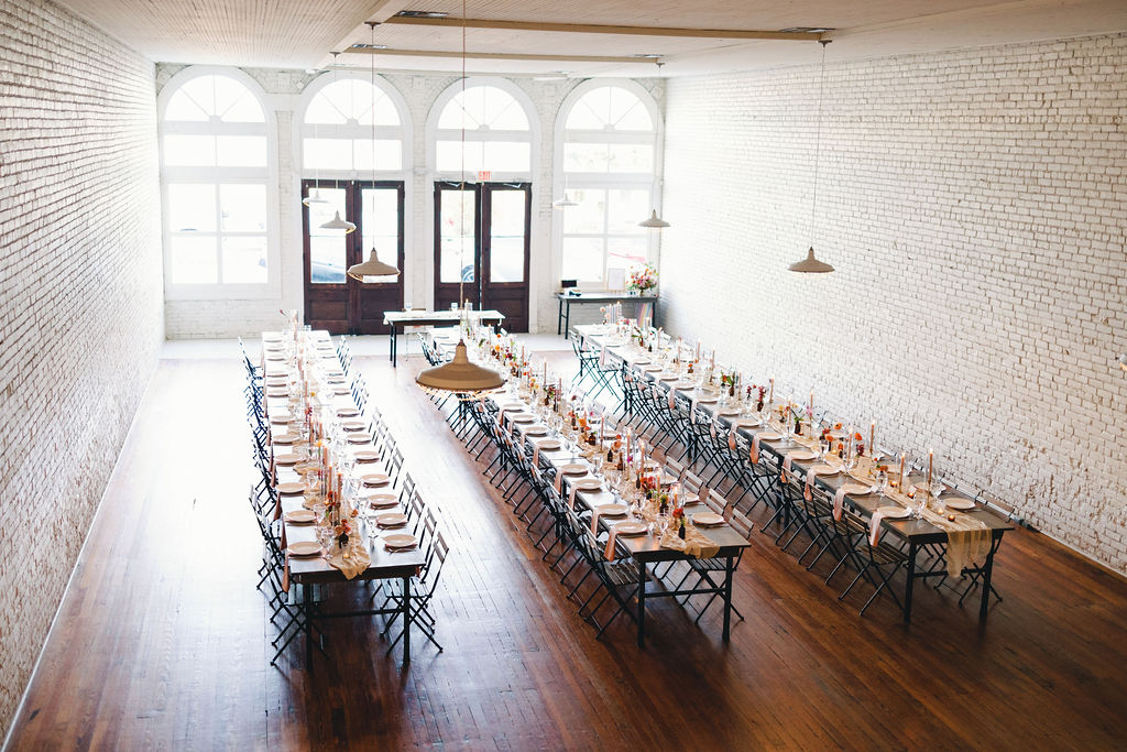 Long tables with place settings and simple floral centerpieces are arranged in a spacious, white-brick room for a One Eleven East wedding, featuring wooden floors and large windows at the far end.