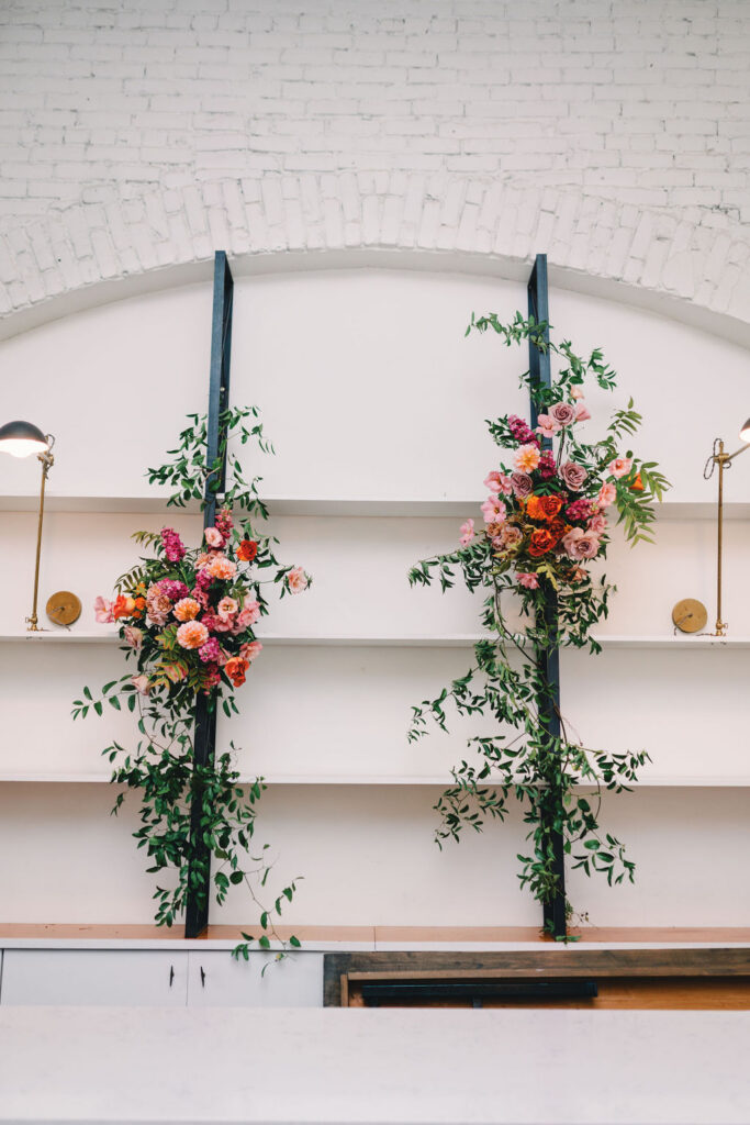 Two floral arrangements with pink, red, and orange flowers and green leaves are mounted on white shelves against a white brick wall at One Eleven East.