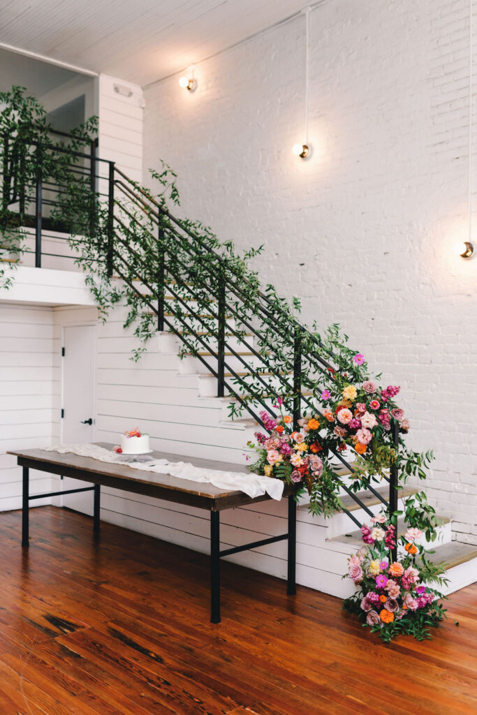 A modern staircase with greenery at One Eleven East is decorated with large, colorful flower arrangements. A long table with a white cloth and small cake stands sits below in a bright room with white walls and wood floors.