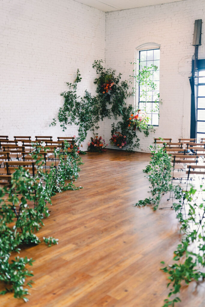 A bright room at One Eleven East features rows of wooden chairs facing forward, green plants lining the aisle, and floral arrangements near a window against a white brick wall.