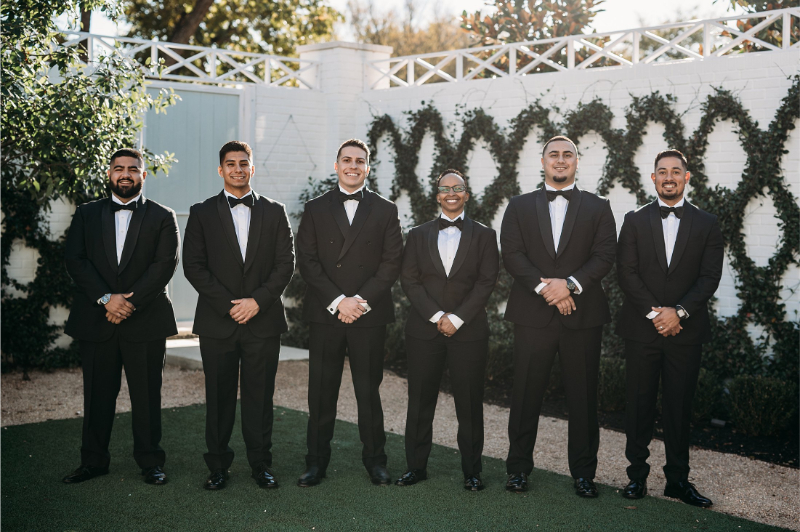 Six men in black tuxedos stand in a row outdoors at Wish Well House, smiling, with greenery and a white fence in the background.