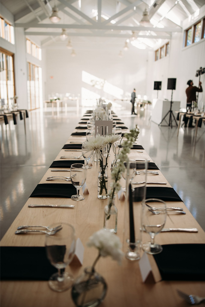 A long, wooden banquet table set with black napkins, glassware, and white flowers graces the bright, spacious event hall of Wish Well House, featuring high ceilings and large windows.