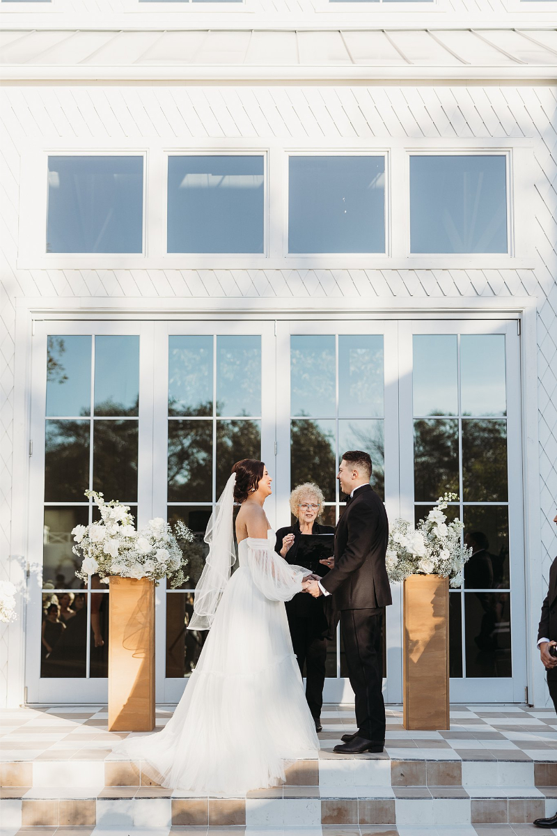 Bride and groom stand facing each other holding hands during a wedding ceremony outdoors in front of the large glass doors at Wish Well House, with an officiant and floral arrangements nearby.