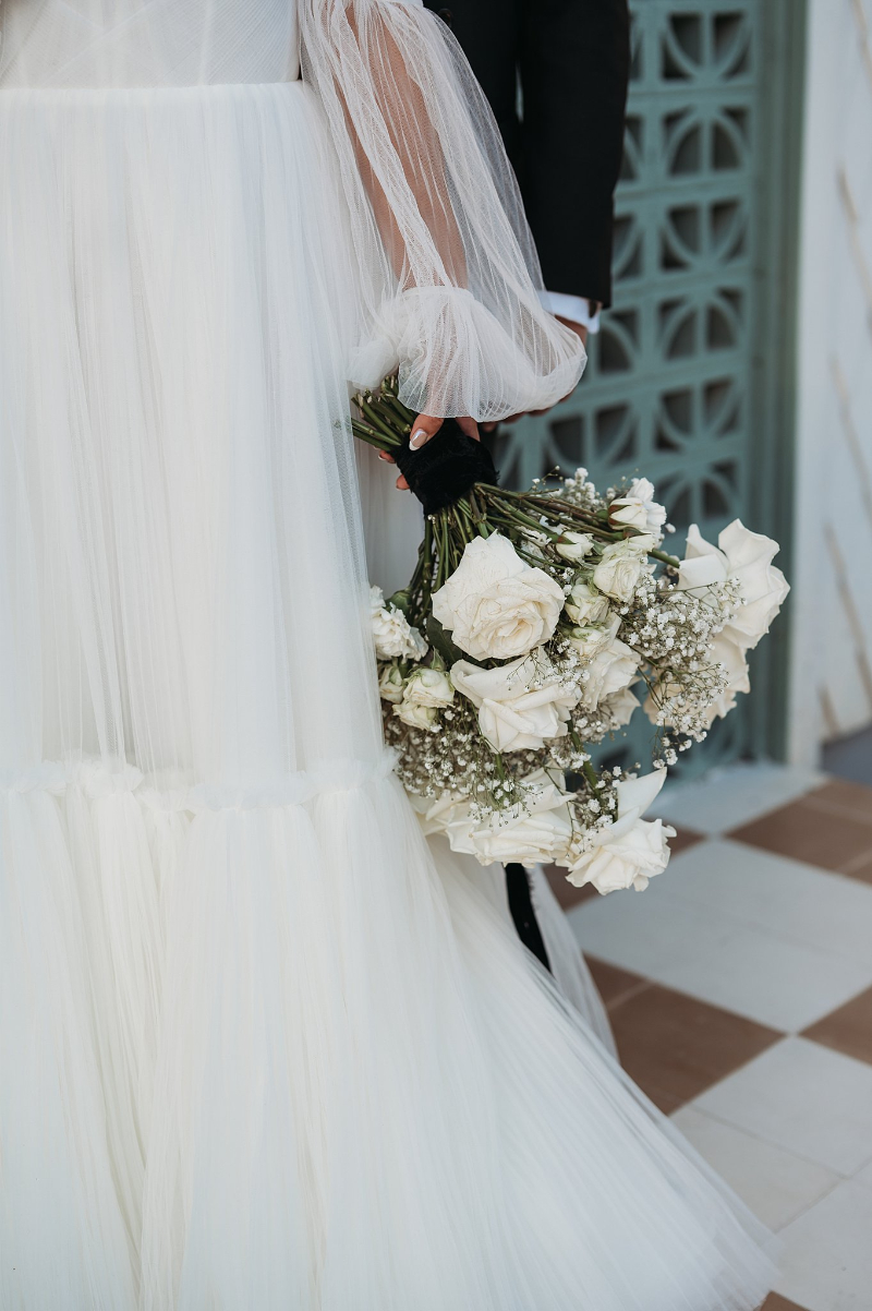 A person in a white wedding dress holds a bouquet of white roses and baby’s breath, pictured at Wish Well House, with part of another person in a dark suit visible beside them.