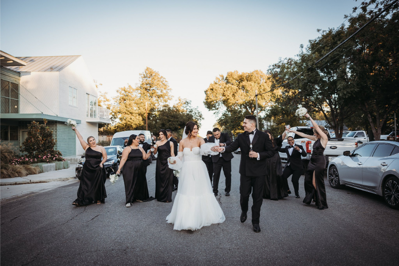 A bride and groom walk joyfully down a street outside Wish Well House, surrounded by cheering bridesmaids and groomsmen dressed in black formalwear.