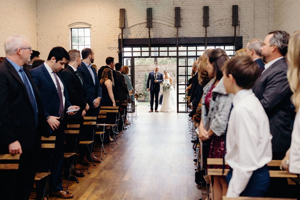 A groom waiting at the altar as guests turn to watch the bride's entrance at a 111 East Wedding.