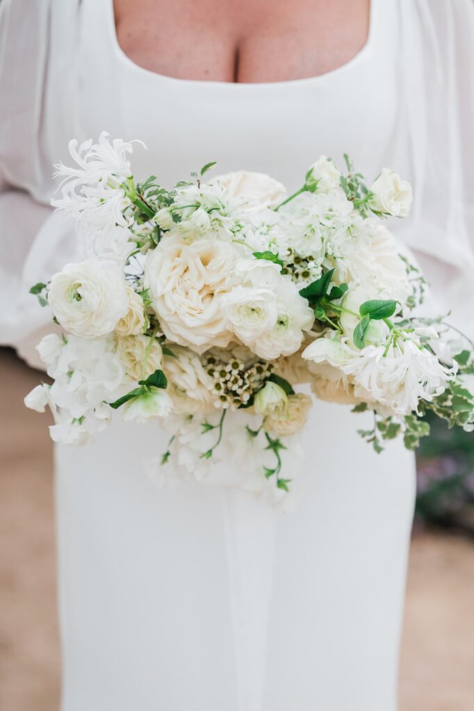 Person in a white dress holding a bouquet of white and cream flowers with green foliage at Two Wishes Ranch.