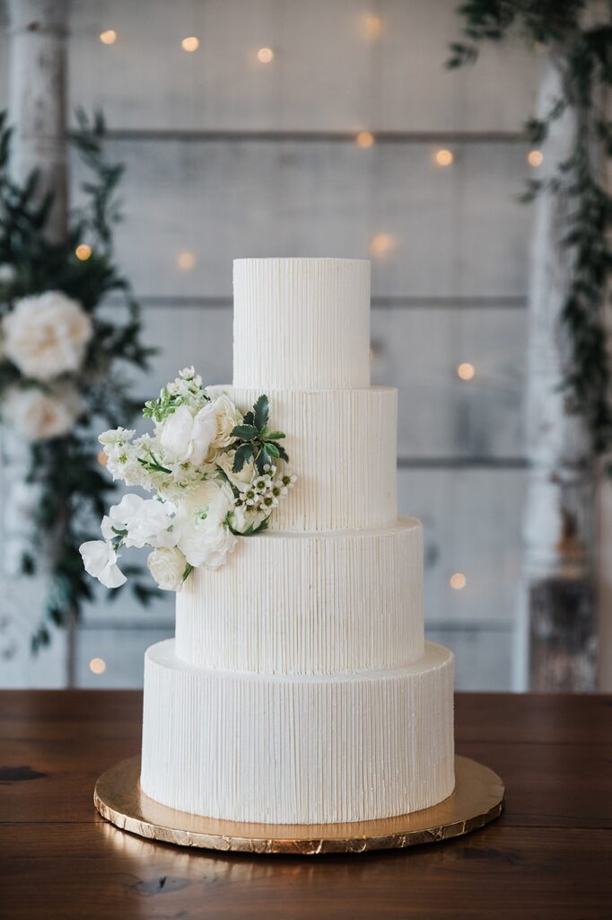 A four-tiered white wedding cake adorned with white flowers is displayed on a wooden table at Two Wishes Ranch. The background features white wooden walls and faint string lights.