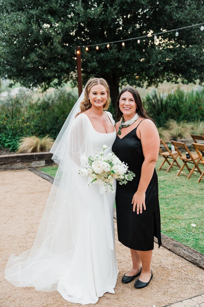 Two women smiling, one in a white bridal gown holding a bouquet and the other in a black dress, standing outdoors with greenery and chairs in the background.