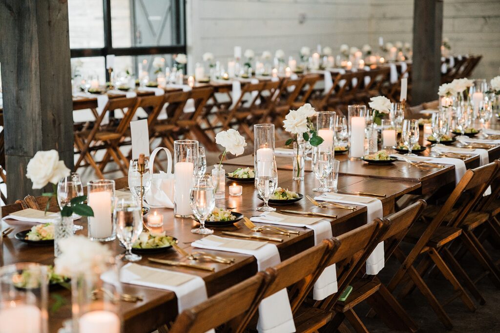 A long banquet table set for an event  featuring plates, glasses, candles, and white flower centerpieces arranged neatly. Rows of similar tables extend into the distance under industrial-style lighting.