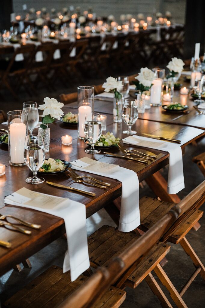 A long banquet table set for a formal event at Two Wishes Ranch, adorned with white flowers, candles, and place settings, including glasses, plates, and cutlery on a wooden table with wooden chairs.