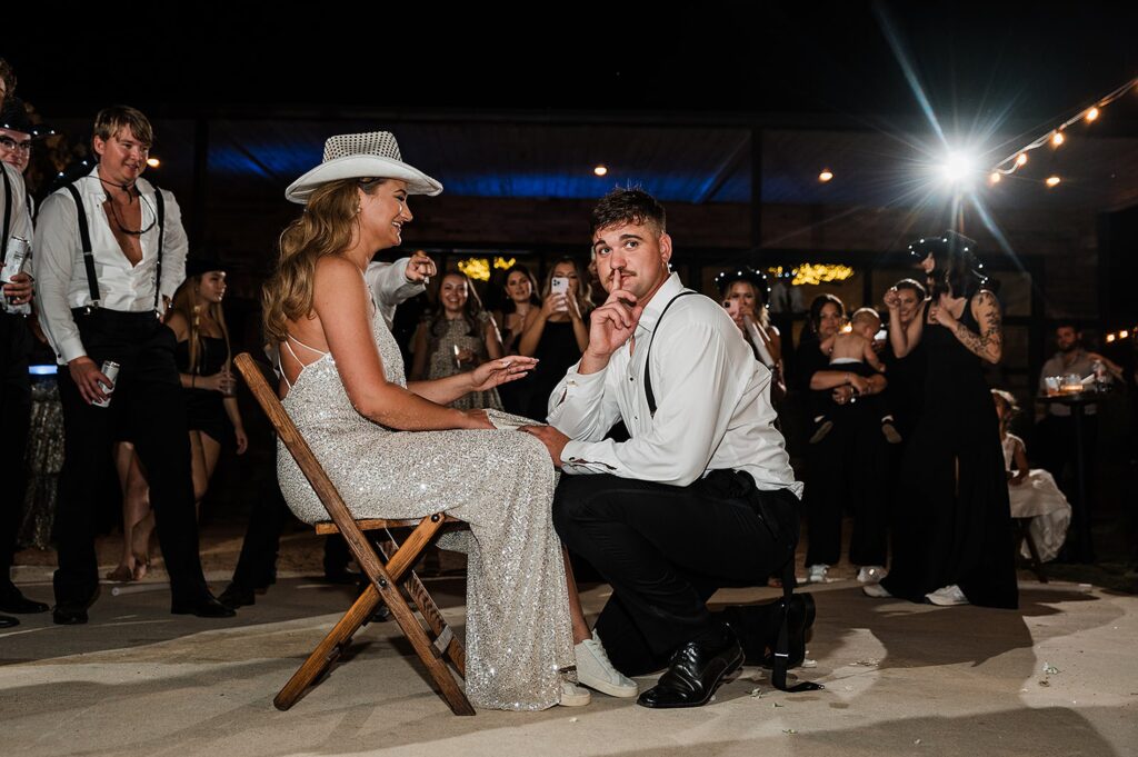 A seated woman in a white dress and cowboy hat is surrounded by a crowd. A man kneels in front of her, holding a finger to his lips. The scene unfolds outdoors at night, with string lights twinkling in the background.