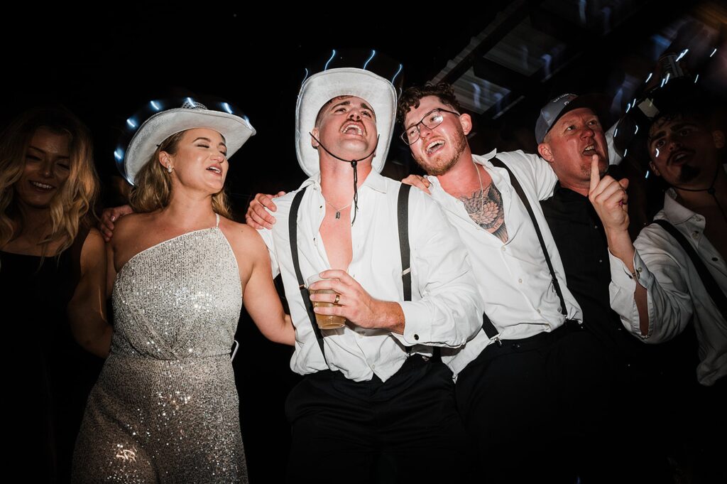 A group of people dressed in light-up cowboy hats and formal attire are laughing and singing together. One person in the center holds a drink. The photo is taken at night.