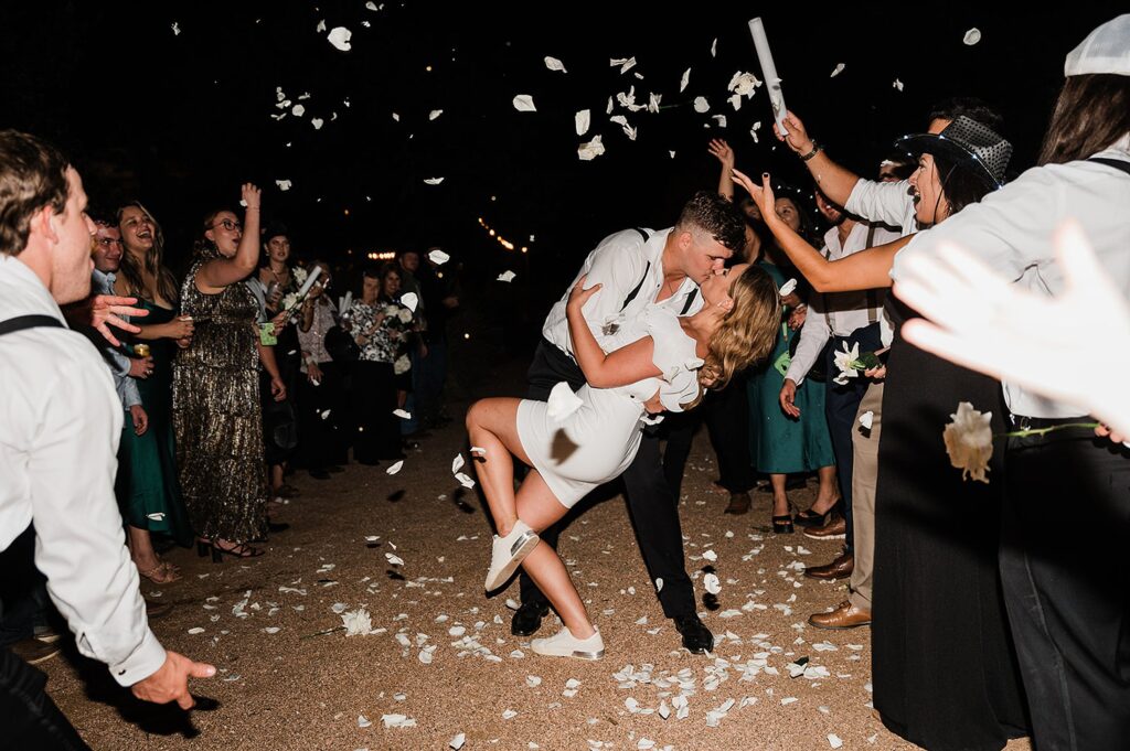 A couple shares a kiss in the center of a crowd at Two Wishes Ranch. Confetti falls around them as people celebrate, cheering and smiling.