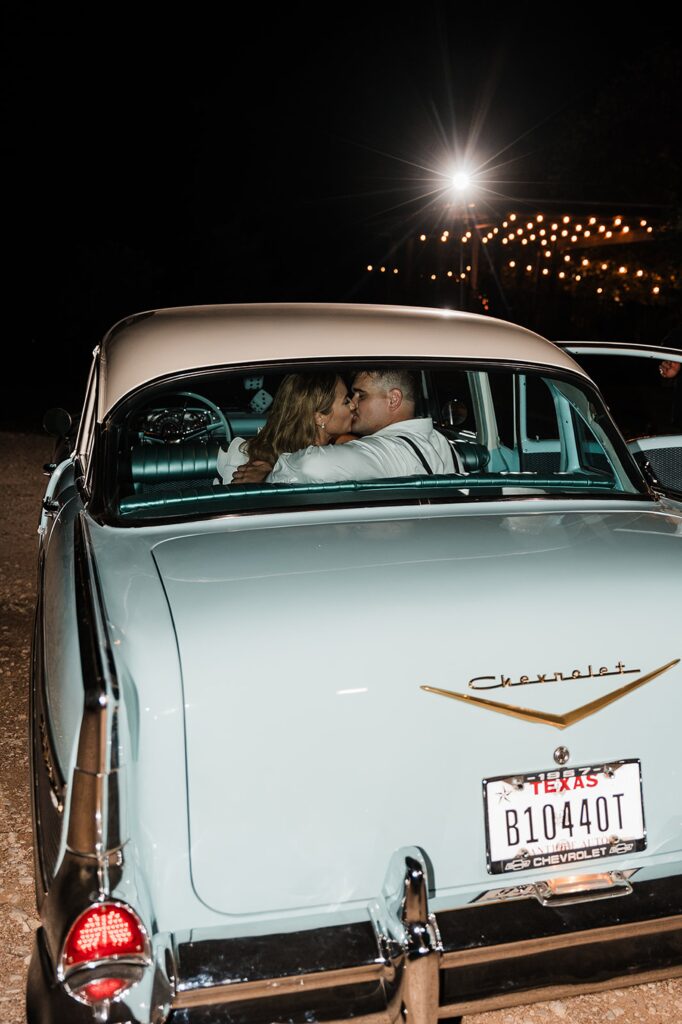 A couple sits in the back seat of a vintage car with a Texas license plate, parked outdoors at night and kissing under the stars. The scene is reminiscent of romantic evenings at Two Wishes Ranch.