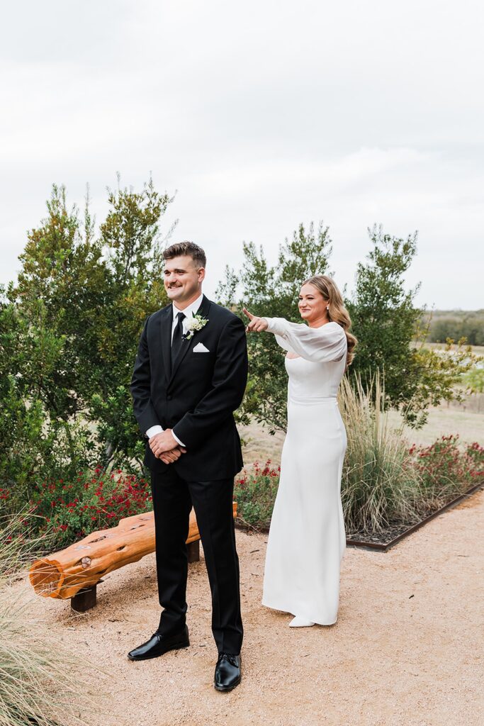 A woman in a white dress is about to surprise a man in a black suit by touching his shoulder from behind at Two Wishes Ranch, surrounded by lush greenery.