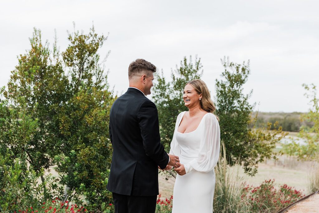 A couple stands outdoors holding hands and smiling at each other, with greenery and an overcast sky in the background.