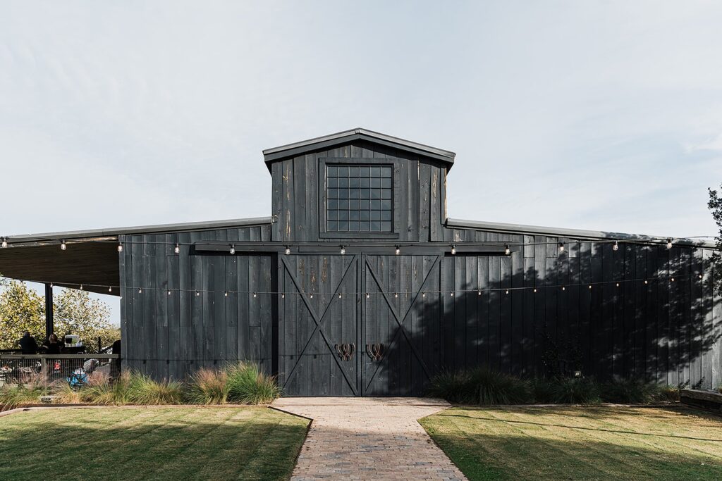 A large, dark gray barn with a central peak and double doors stands at the end of a brick path, surrounded by grass and illuminated by string lights. This picturesque scene is part of Two Wishes Ranch.