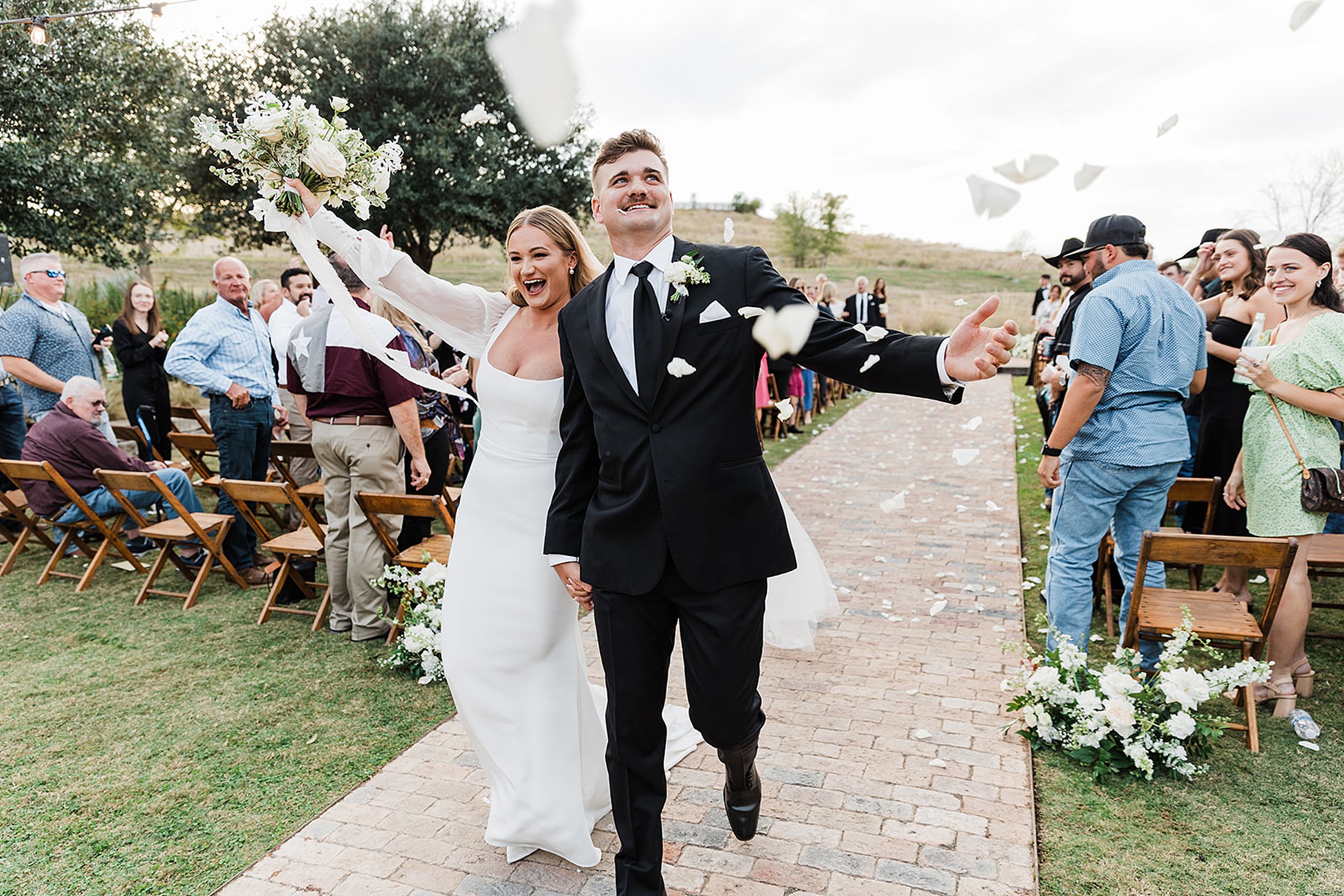 A newlywed couple walks down the aisle smiling and holding hands at Two Wishes Ranch, as guests toss flower petals in celebration.