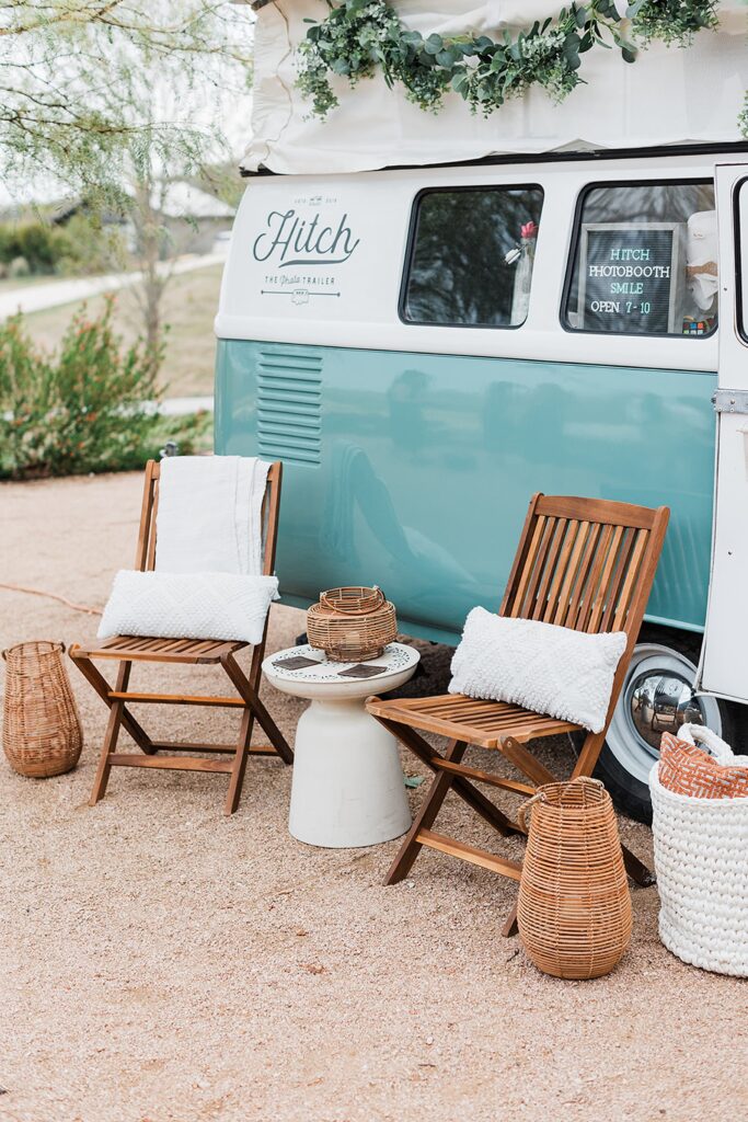 Two wooden chairs with white cushions sit next to a light blue and white vintage van at Two Wishes Ranch. Decorative baskets are placed around, and a white blanket is draped over one chair. Lush greenery is on top of the van.