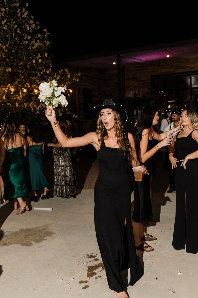 A woman in a black dress holding a white flower bouquet raises her arm while standing outside at night at Two Wishes Ranch, surrounded by people gathered in formal attire.