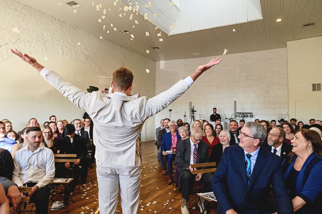 A person in a light suit stands with arms raised, facing a seated audience in a brightly lit room as paper confetti falls from above .The audience appears engaged and smiling, enjoying the celebration.