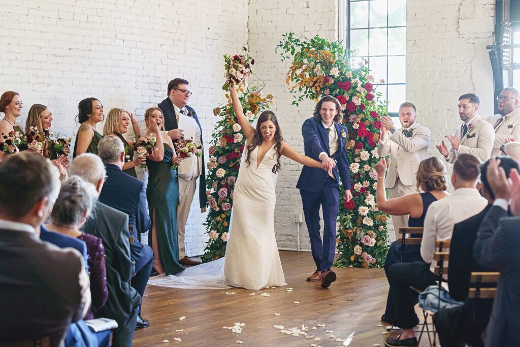 A newlywed couple joyfully exits down the aisle while guests applaud at their Austin Winter Wedding. Bridesmaids and groomsmen stand on either side, and floral arrangements adorn the background, capturing the magic of a Winter Wedding at One Eleven East.