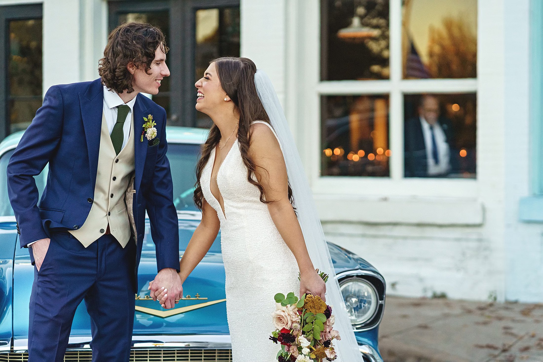 A bride in a white gown and a groom in a blue suit hold hands and smile at each other in front of a blue vintage car, perfectly capturing the charm of an Austin Winter Wedding at One Eleven East.