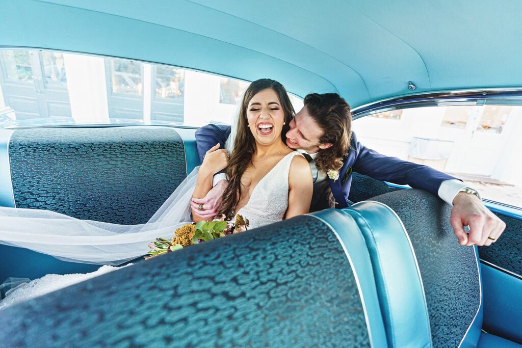 A bride and groom share a joyful moment in the backseat of a vintage car during their Austin Winter Wedding; the groom playfully nuzzles the bride, causing her to laugh.