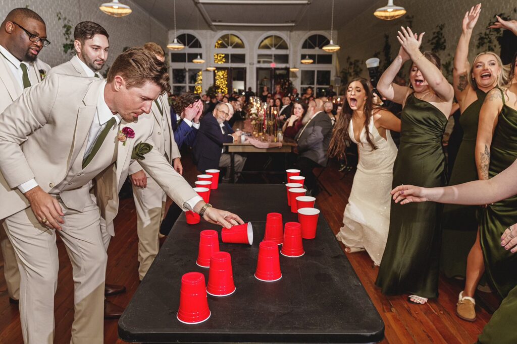 A group of people dressed formally are playing a game with red cups on a table. Some are actively participating, while others are cheering and watching.