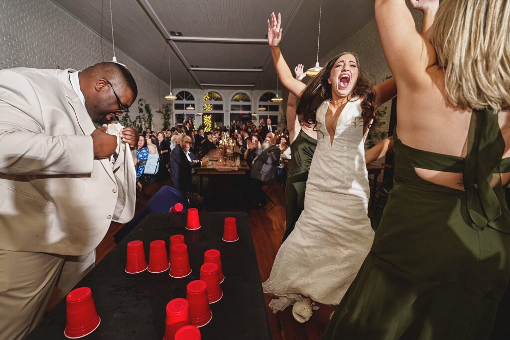 A bride in a white dress and bridesmaids in green dresses enthusiastically play a game with red cups on a long table, while a man in a light-colored suit nearby looks down, focusing on his action.