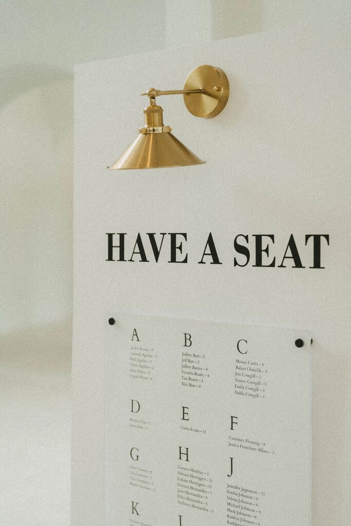 A brass wall lamp is mounted above a sign reading "HAVE A SEAT". Below the sign is a seating chart with names organized alphabetically.