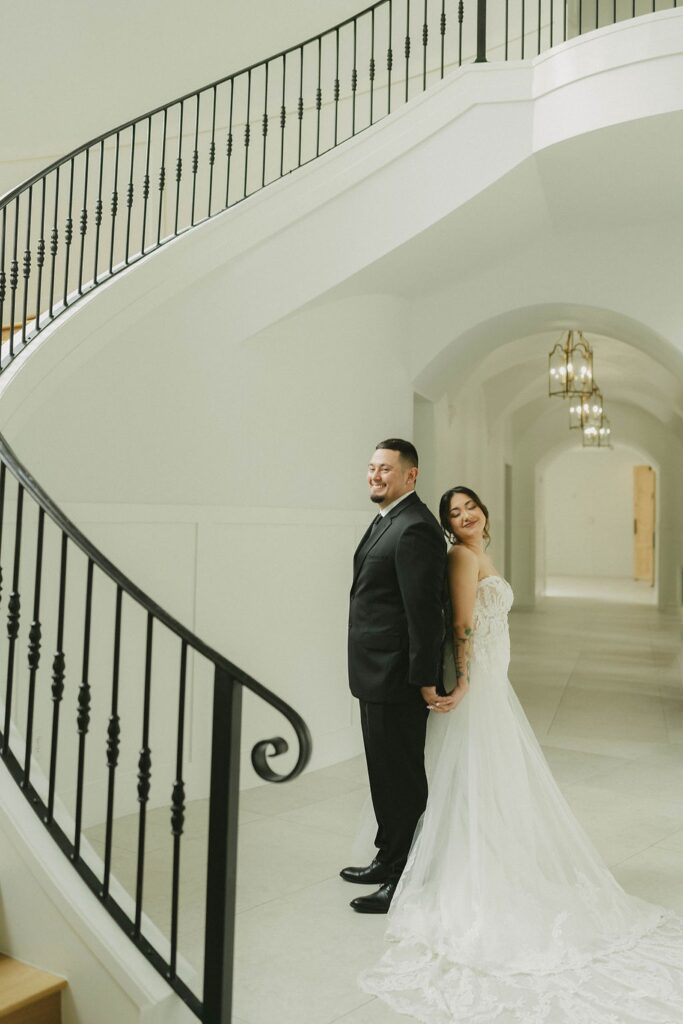 A bride and groom stand back-to-back holding hands, smiling, in front of a white, curved staircase with black railings at The Preserve at Canyon Lake. The bride wears a white gown and the groom a black suit.