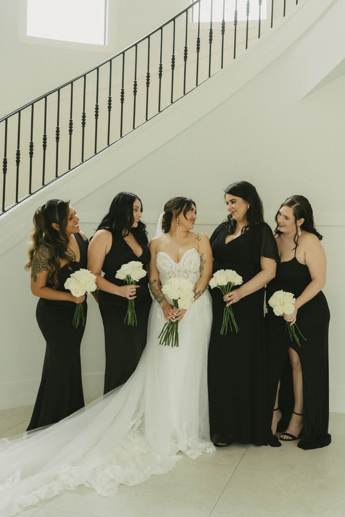 A bride in a white gown stands beside four bridesmaids in black dresses, all holding white bouquets, posing in front of a modern staircase at The Preserve at Canyon Lake.