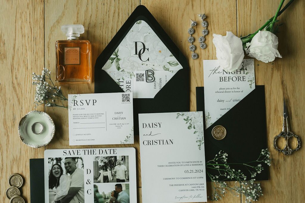 A collection of wedding stationery lies on a wooden surface, including invitations, RSVP cards, a "Save the Date" card for The Preserve at Canyon Lake, a small perfume bottle, white flowers, scissors, and a wax seal stamp.