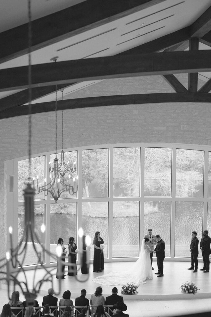 A wedding ceremony taking place in a spacious hall at The Preserve at Canyon Lake with large windows and a high ceiling. The bride and groom stand at the altar while the officiant speaks and the guests are seated watching.