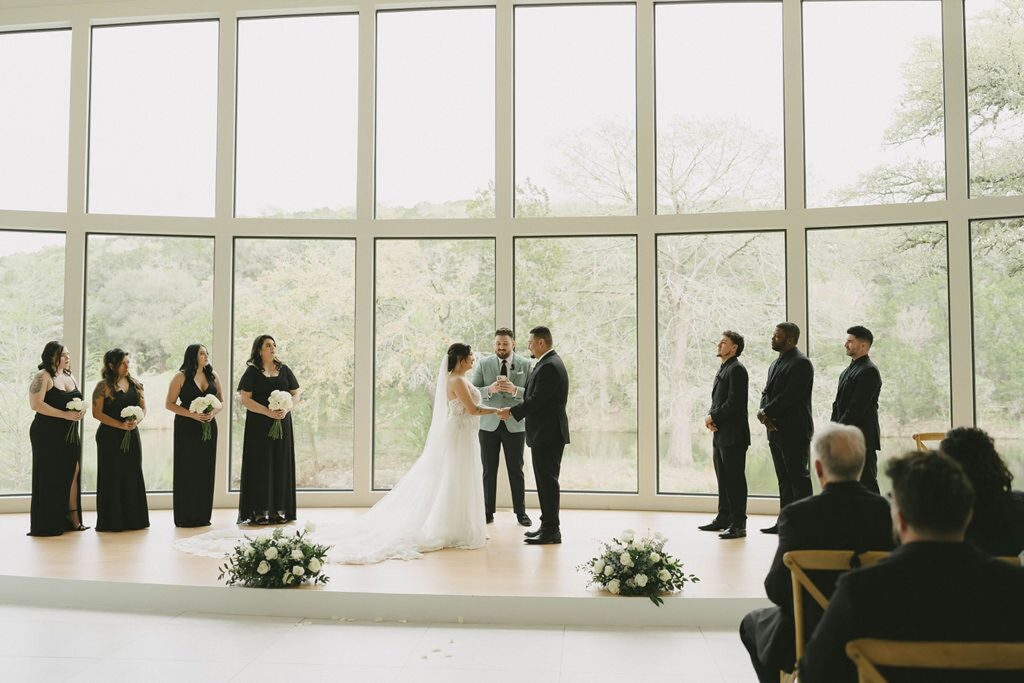 A couple exchanges vows at a wedding ceremony in front of large windows. The bride and groom are flanked by their bridal party, with guests seated in the foreground.