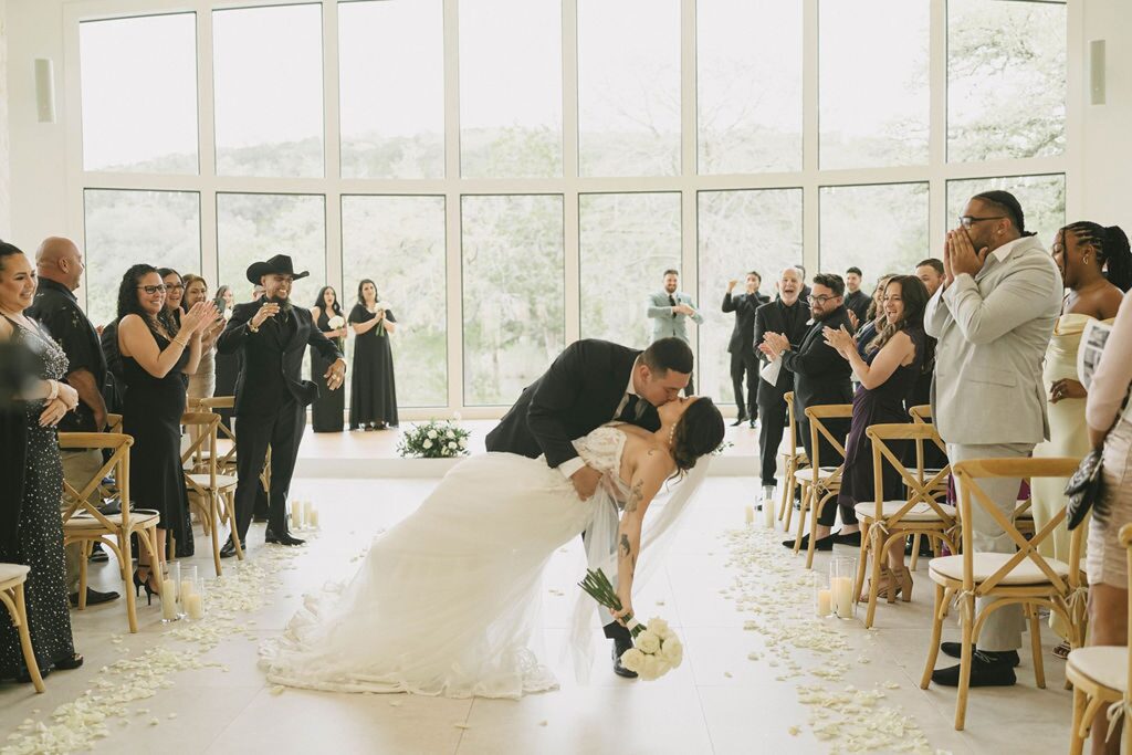 A newlywed couple shares a kiss at the end of the wedding aisle at The Preserve at Canyon Lake, surrounded by applauding guests in a bright venue with large windows and white rose petals scattered on the floor.