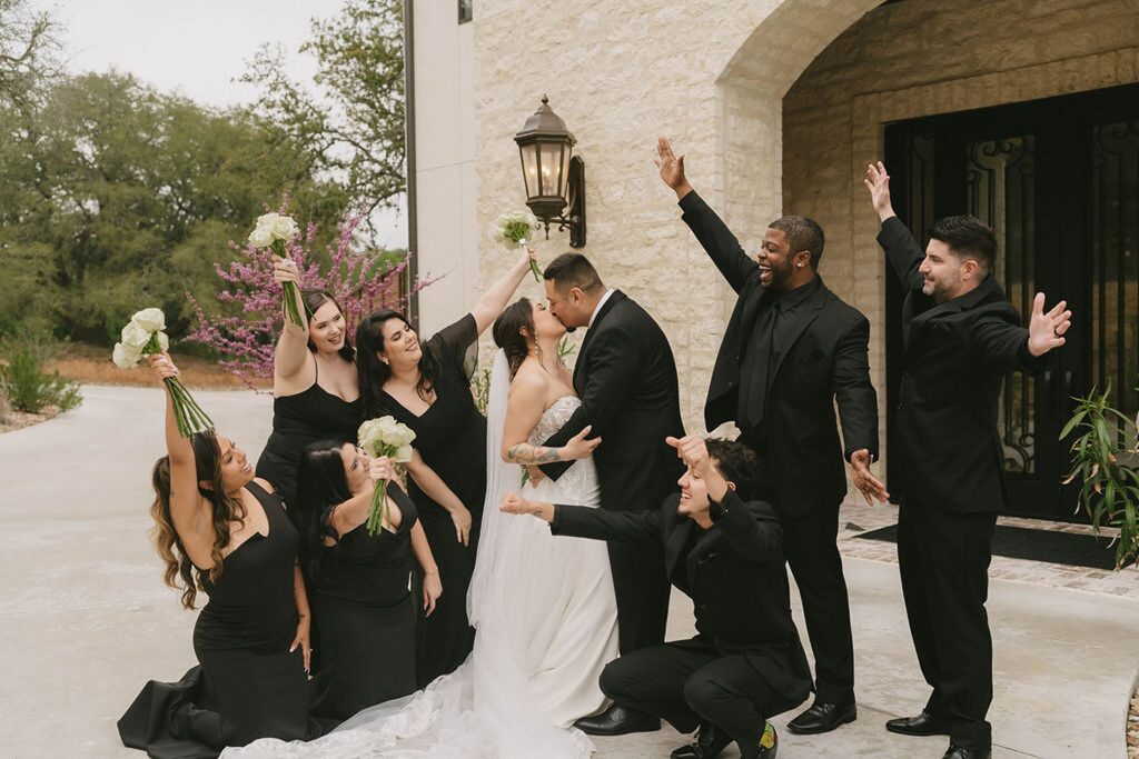 A newlywed couple kisses at The Preserve at Canyon Lake, surrounded by six wedding party members in black outfits, all cheering and celebrating with raised hands and bouquets.